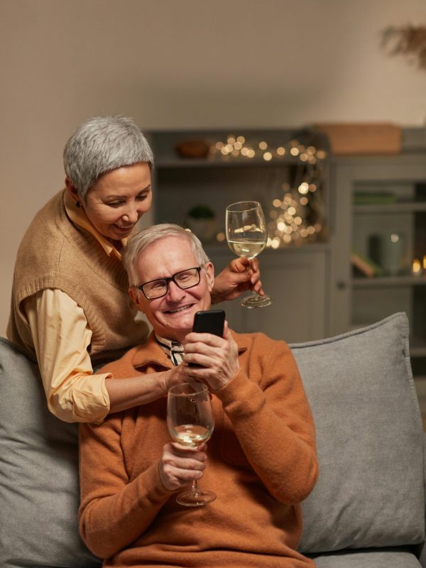 Senior couple smiling with wine glasses, enjoying leisure time and technology indoors.